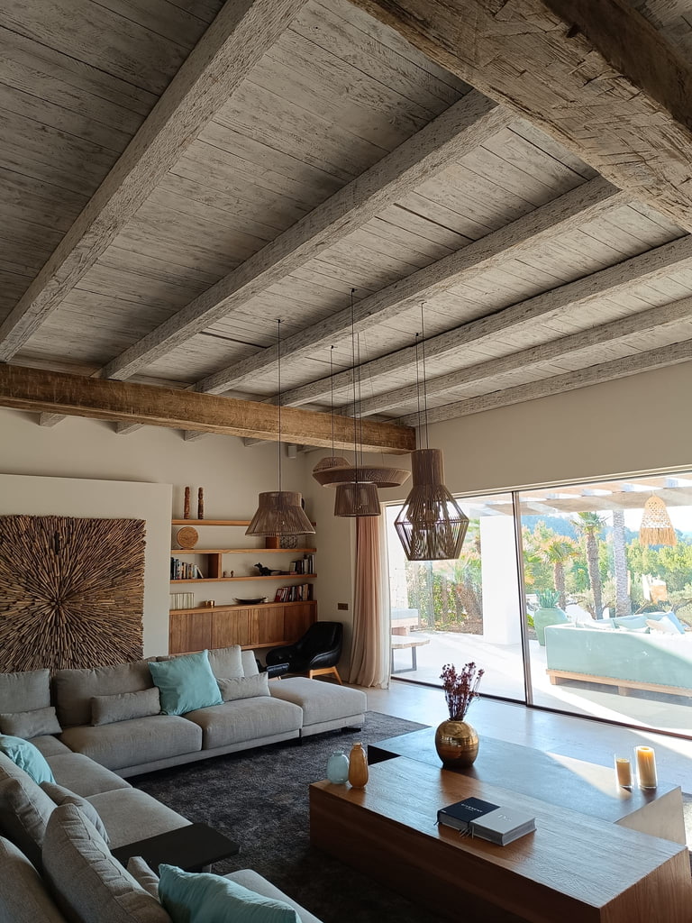 Salón de villa en Ibiza con techo de vigas de madera, estanterías a medida y panel decorativo de madera